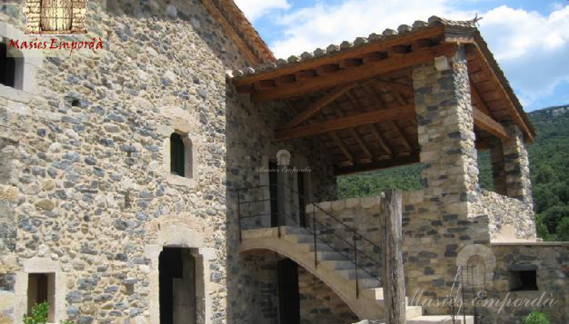 Vista del conjunto constructivo principal de la masía desde el patio interior con cubierto anexo a la fachada de la casa, a dos aguas con pilares de piedra, tejado de teja antigua y vigas de madera.  