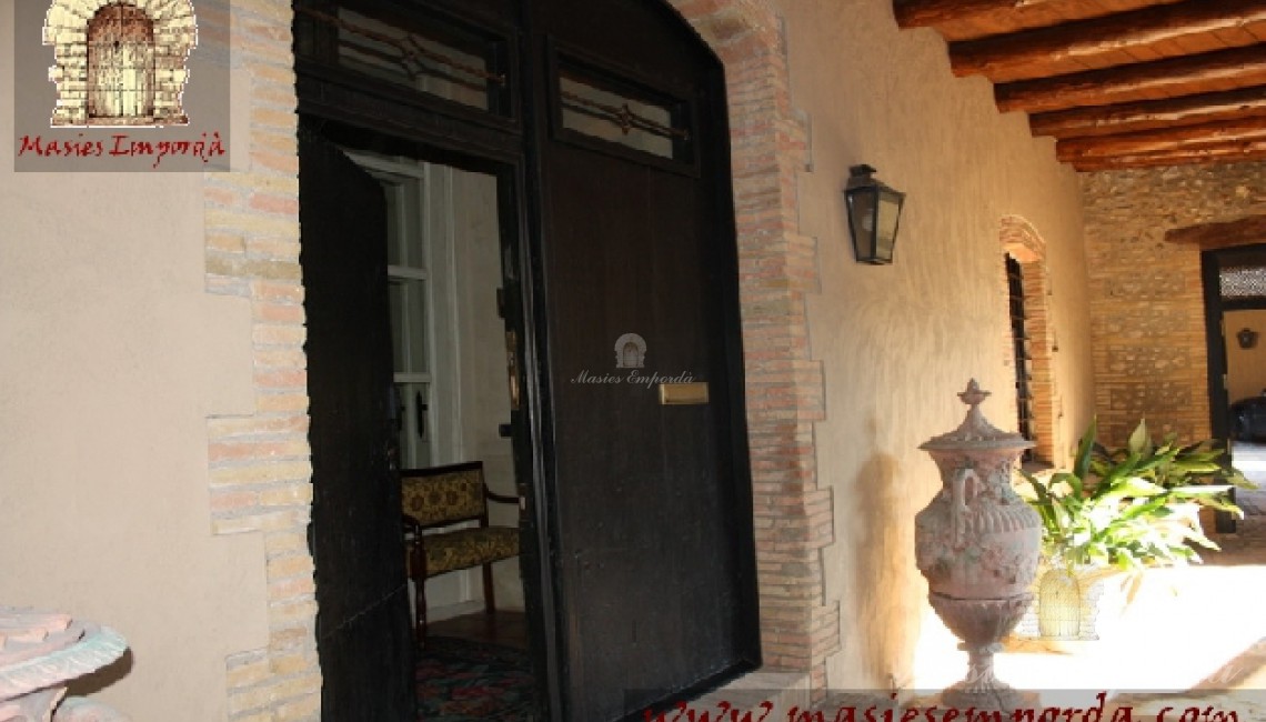 Gran puerta principal de madera y contra puerta interior también de madera y acristalada con el porche corrido en fachada con vigas de madera  