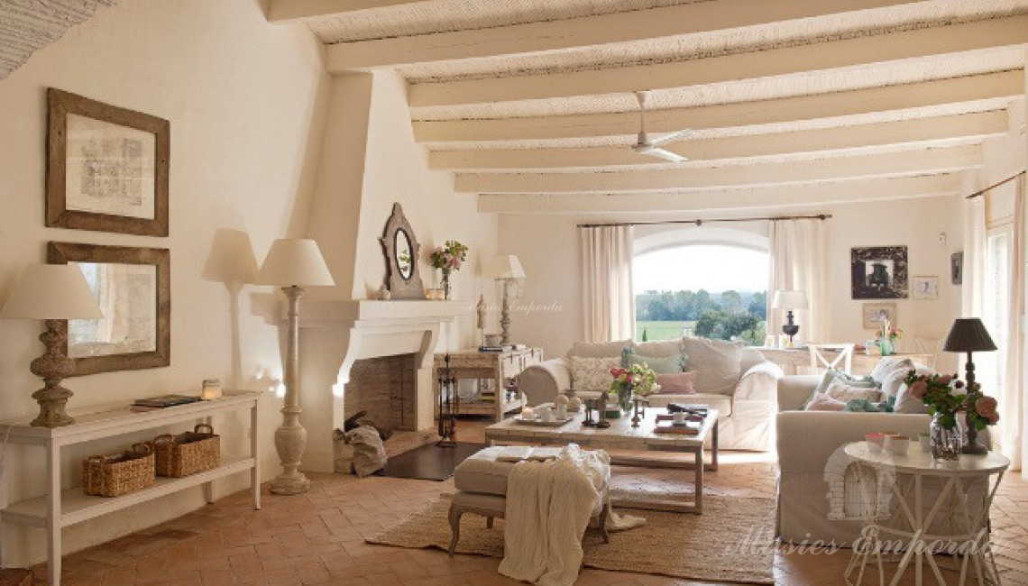 Salón de estar con vigas de madera vista y pintadas como el resto de la sala con chimenea en el centro de esta y un ventanal al fondo con vistas a los campos cercanos 